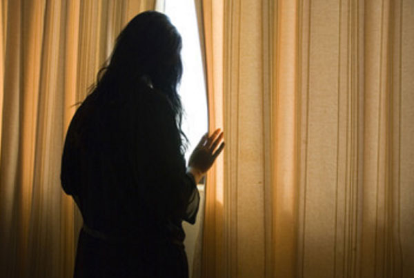 Женщина стоит возле зашторенного окна. С опаской выглядывает на улицу