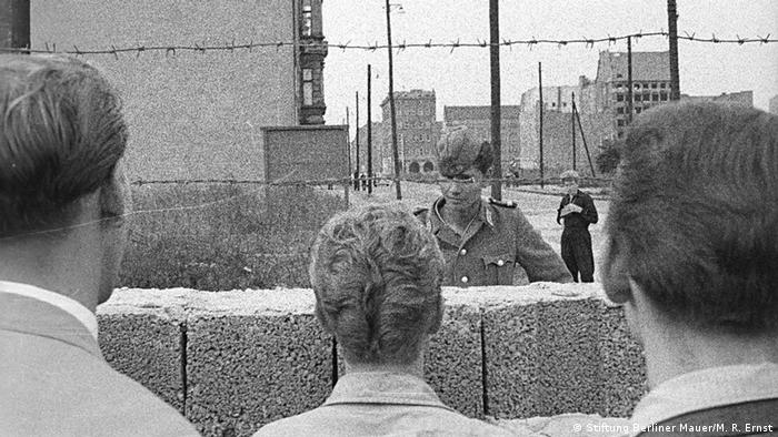 A solider stands on one side of the Berlin wall as it is under construction while people look on (Stiftung Berliner Mauer/M. R. Ernst)