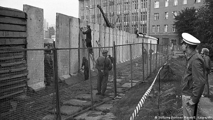 Workers patch the cracks between the concrete segements as the new Berlin Wall gets installed while a West policeman looks on (Stiftung Berliner Mauer/E. Kasperski)